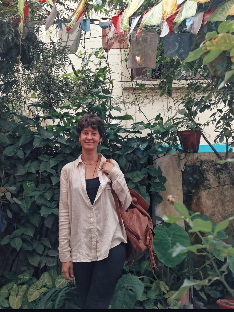 Ich stehe vor einer Hauswand voller Pflanzen und trage mein helles Leinenhemd und meinen braunen Rucksack über einer Schulter